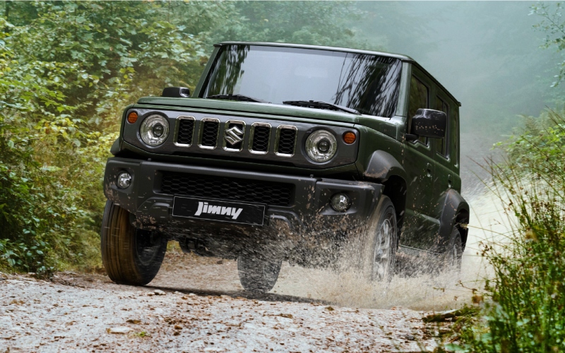 Carro 4x4 Suzuki Jimny verde desplazándose por un camino de tierra en un bosque, salpicado de agua.